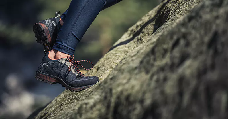 Een persoon met donkere wandelschoenen aan beklimt buiten een rotsachtig oppervlak; alleen de onderbenen en voeten zijn zichtbaar. De scène doet denken aan wandelen of een buitenactiviteit in de natuur.